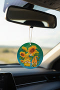 A round car air freshener hanging from a rearview mirror inside a vehicle. The disc features a colorful illustration of sunflowers, bees, and a small orange cat on a teal background.