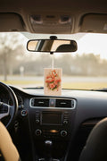 A rectangular car air freshener or hanging ornament with a pink flamingo and floral design, suspended from the rearview mirror inside a car.