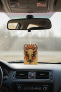 A rectangular car air freshener with a detailed skull and floral design hanging from a suction cup mount on the rearview mirror inside a car.