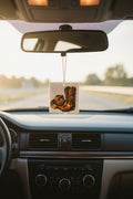 A square car air freshener with a printed cowboy boots design hangs from the rearview mirror inside a car, with the road and dashboard visible in the background.