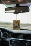 A rectangular car air freshener with a colorful floral print hangs from the rearview mirror inside a car dashboard.