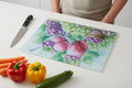 A rectangular glass cutting board with a colorful fruit and wine illustration sits on a white table. A person in a beige top is partially visible behind it, with a knife, carrots, red bell pepper, yellow bell pepper, and zucchini arranged around it.